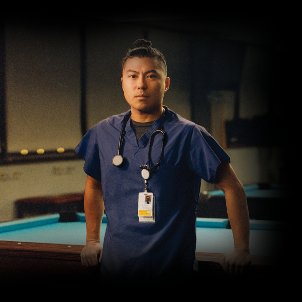 Dr. Derek Wong, poses in a pool hall in his scrubs looking at the camera.